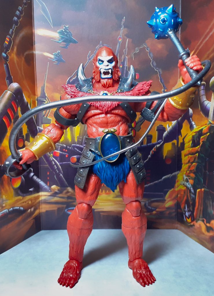 A photo of the New Eternia version of Beast Man. This figure is standing and looking right at the viewer. It is largely orange in colour and moulded to look furry and hairy. The figure is brandishing a whip in its right hand and holding up a spiked mace in its left.