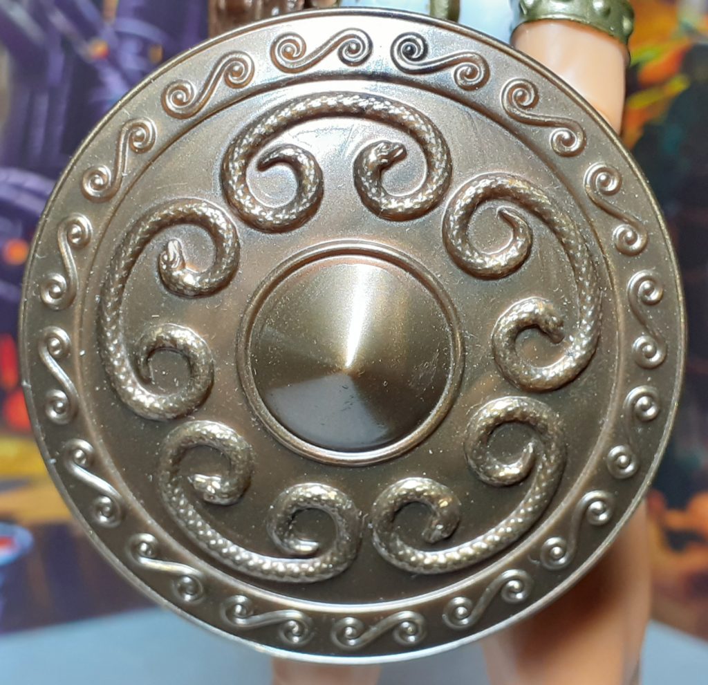 A close up look at the shield for New Eternia Teela. This is the front of the shield which is moulded in that shiny bronze coloured plastic. The shield has raised details on it including curled snakes and a pointy central area.