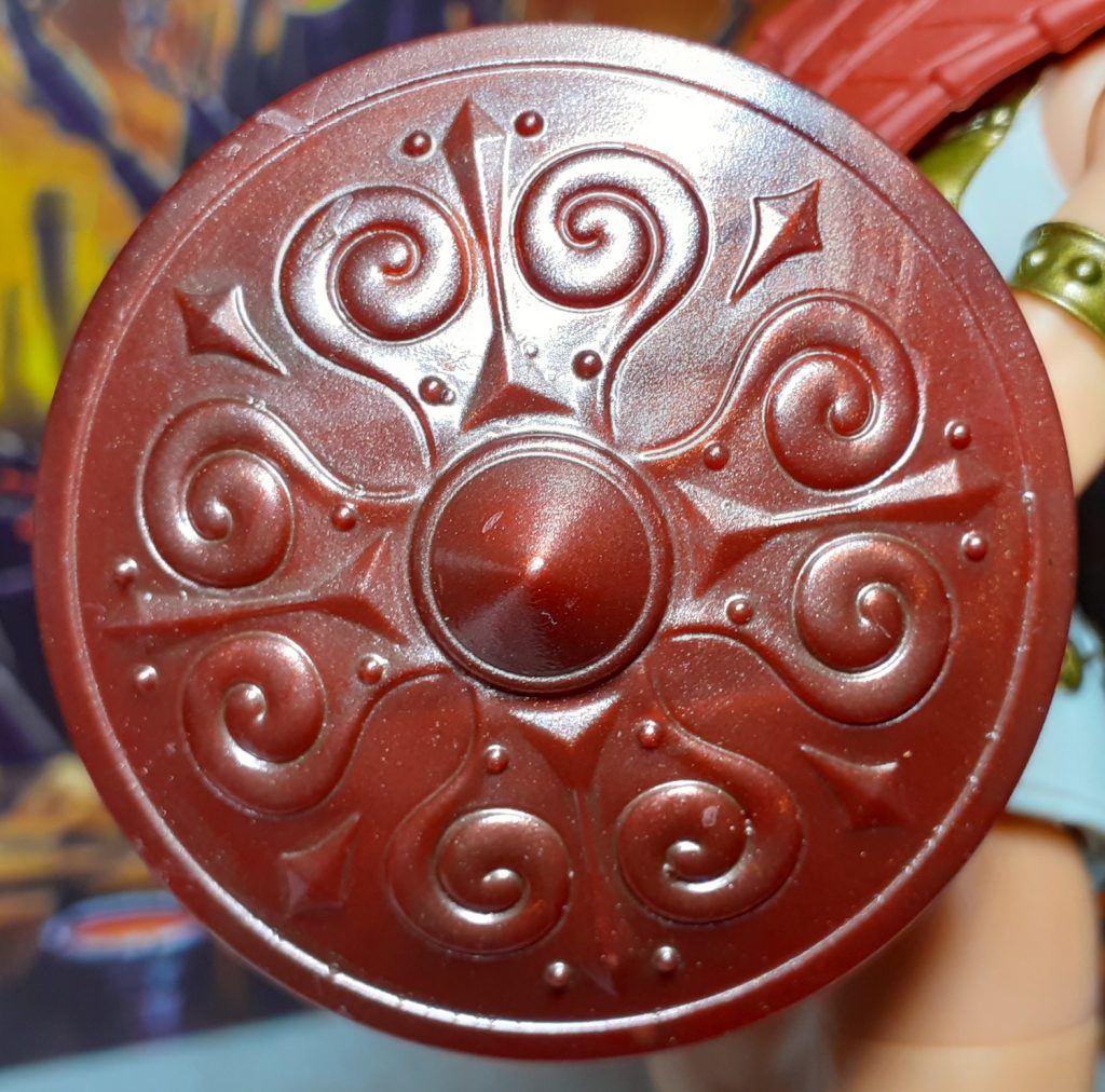 A close up photo of the shield for Vintage Collection Teela. The front of the shield has a raised pointy central area along with curving designs around it. The shield is a reddish brown again but has a metallic sheen to it.