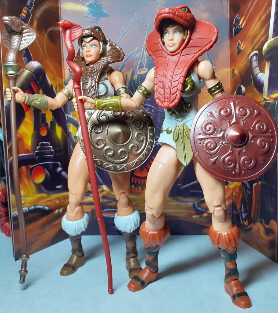 The New Eternia Teela figure on the left with the Vintage Collection Teela on the right. Both figures are standing straight and looking off to the viewer's left. Both figures are wearing their versions of the snake armour, staff and shield.