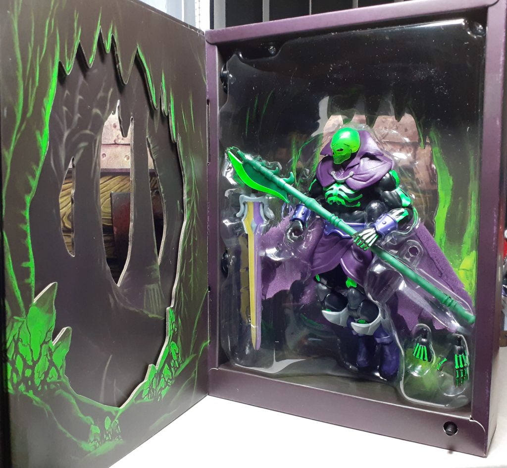 In this photo, the first page of the box is opened and the figure of Scare Glow is finally fully revealed. It's fitted into clear plastic packaging so that it looks like the figure is floating slightly off the floor. The figure is holding a halberd weapon accessory. To the left in the packaging is the Sword of Power while on the right is a pair of spare hands.
