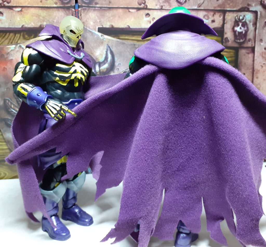 A photo of the back of the SDCC Scare Glow figure showing the cape. This is made from a purple felt material and has a wire running down the inside of both the left and right edges. This means the cape can be moved a posed by bending the wire. As here, the standard release Scare Glow is holding the left edge of the other figure's cape.