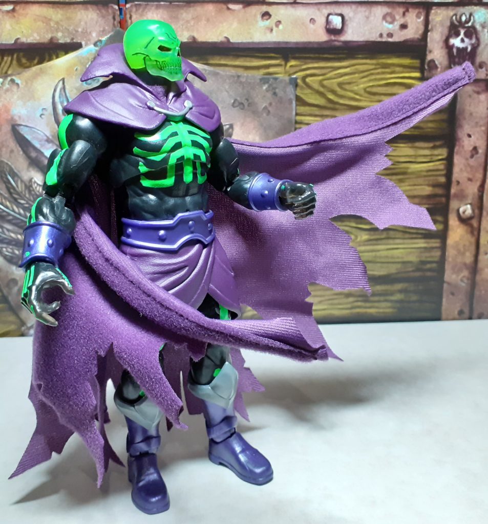 In this photo, the SDCC Scare Glow is standing on the left of the photo, looking to the right. Its cape has been posed to look as though a wind is blowing about and in front of the figure.