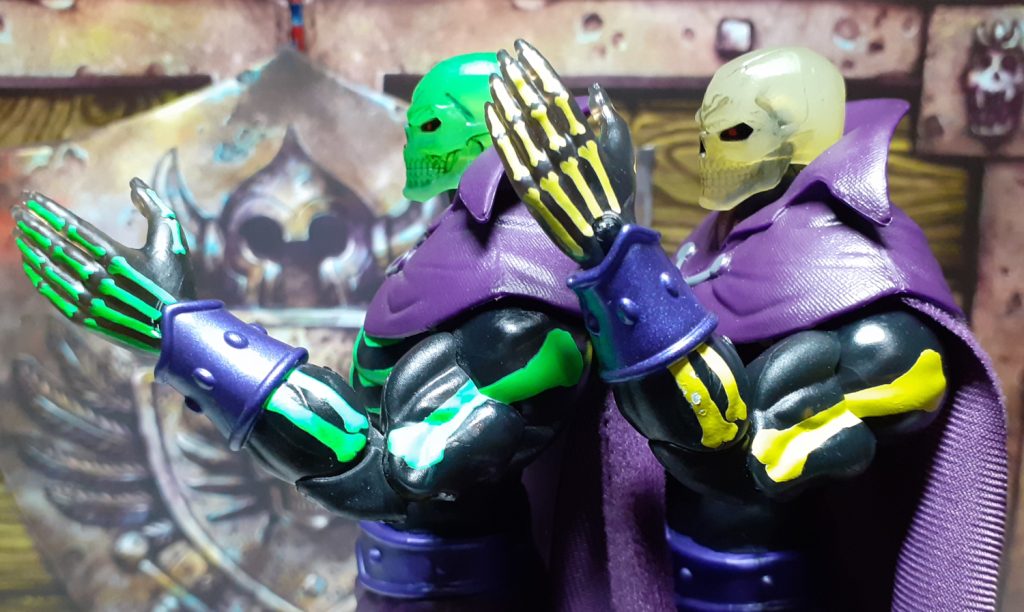 The two Scare Glow figures standing one behind the other. The SDCC version is on the left of the photo while the retail version is on the right. Both figures are facing to the viewer's left and are holding up their left arms. This gives a good comparison of the "bones" painted on their arms and hands.