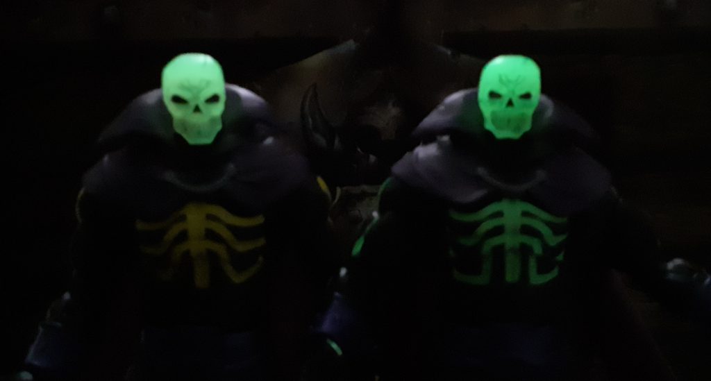 The two Scare Glow figures side by side in the dark. Both figures have glow in the dark heads which can be seen here although the photo is rather blurry. The SDCC Scare Glow's skull head is on the right and glows with more of a lime green.