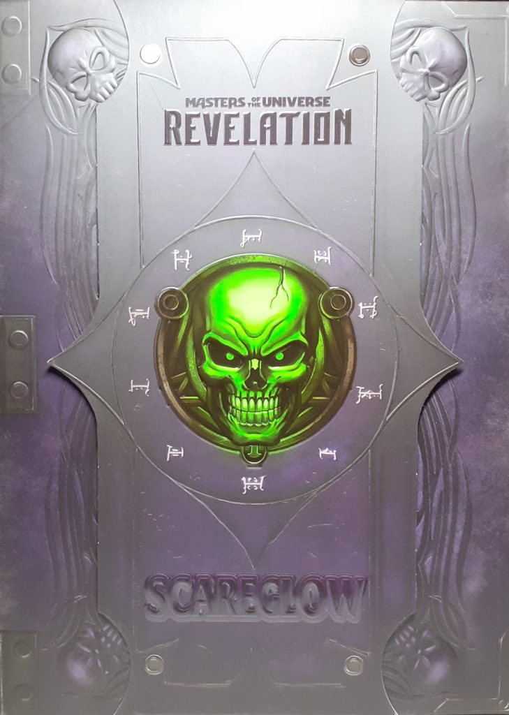 The front of the actual display box for Scare Glow. It has a card board slip over it with Scare Glow's bright green skull face on it. Behind the slip it can be made out that the box's design looks like an ancient book.