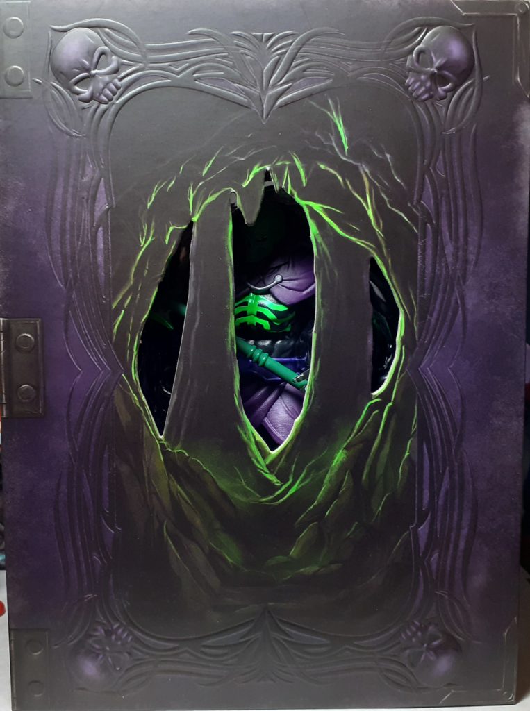 The front of the display box with the cardboard slip removed. Although this looks like the cover to an old book, there are holes cut out in the middle giving the impression of looking through a cave. In the middle of which the figure of Scare Glow can be made out.