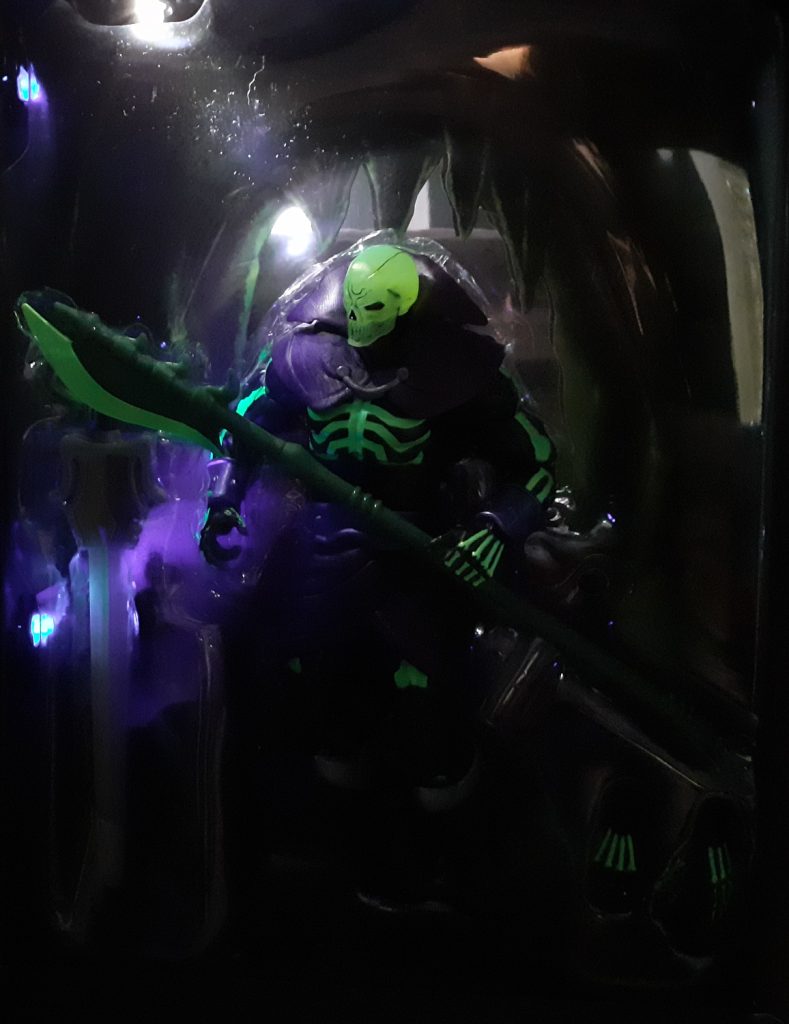 A photo of the Scare Glow figure in its box in the dark. To the top left is a small light angled towards the figure while to the lower left is another light. This second light is a black light that makes the day glow paint on the figure glow a livid green.