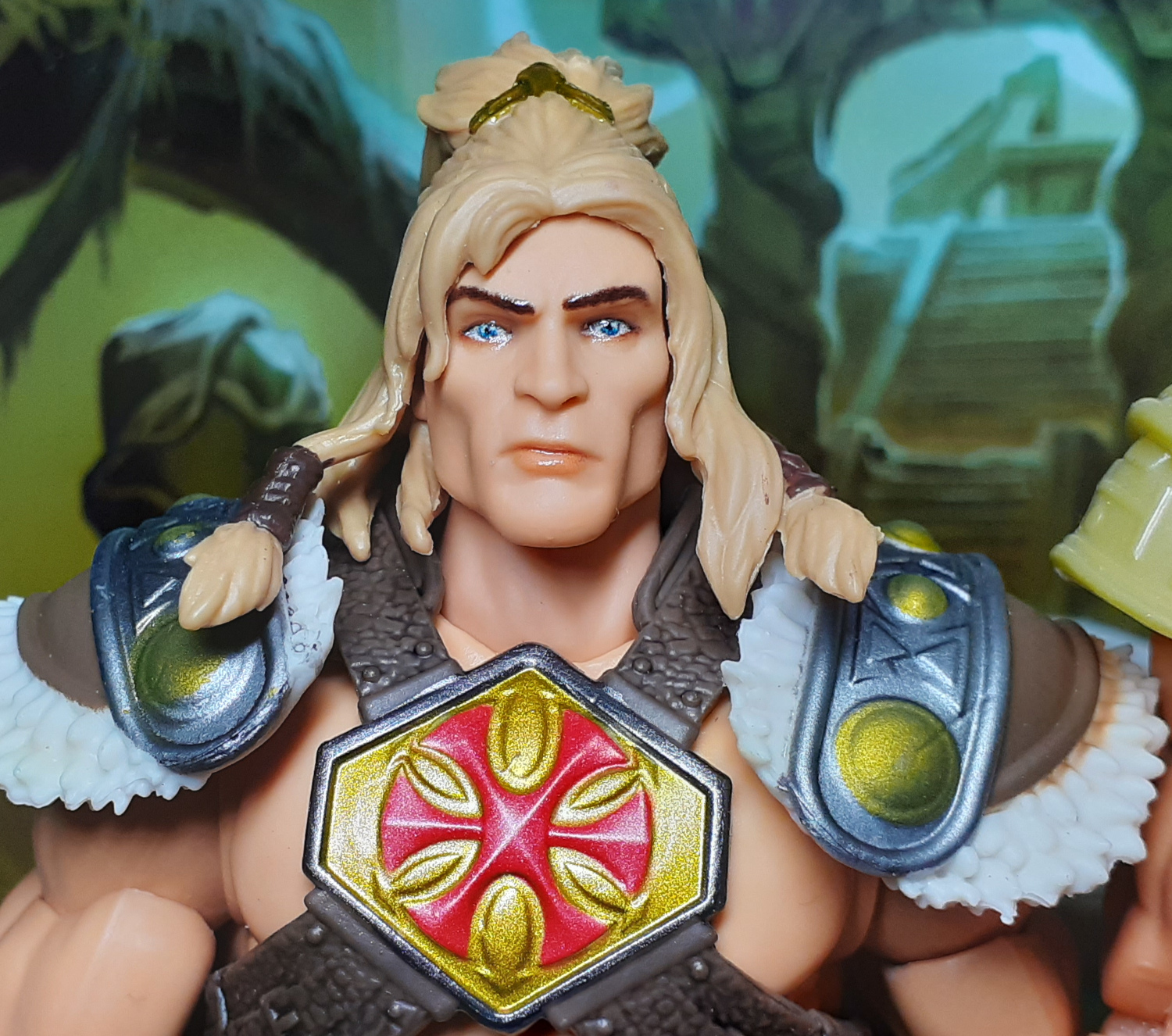 A head and shoulders photo of the King Grayskull figure. The figure has long blonde hair gathered up into a bun on top of its head. It also has hair braids handing down over the large pieces of shoulder armour. The face sports a very square jawed chiselled look. Over the chest the figure has its signature emblem of a red six pointed cross or asterisk on a gold background.