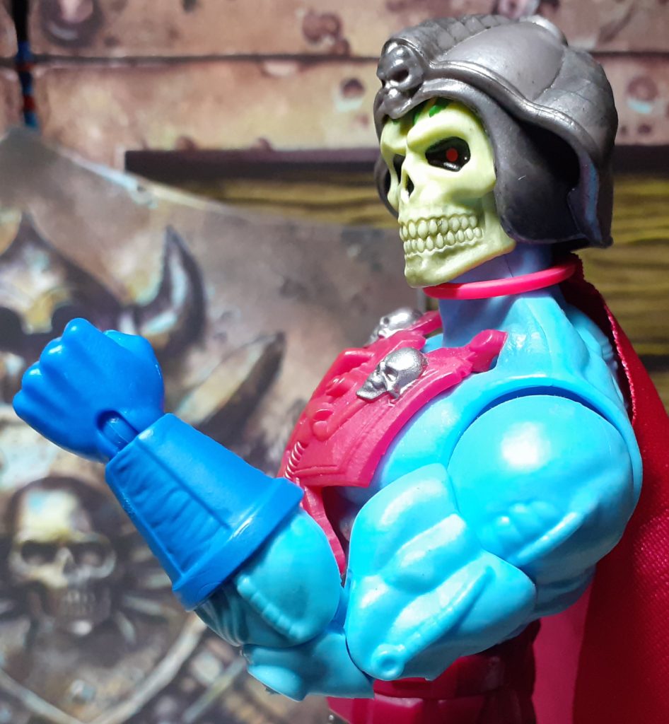 A side view of the top half of New Adventures Skeletor. The figure is holding up its left arm, bent at the elbow as though it is flexing its biceps. The skin tone is a light blue while the hand and lower arm are covered by a darker blue gauntlet. Visible on the arm are some moulded on wires and tubes, giving the impression of being some sort of cybernetic implants.
