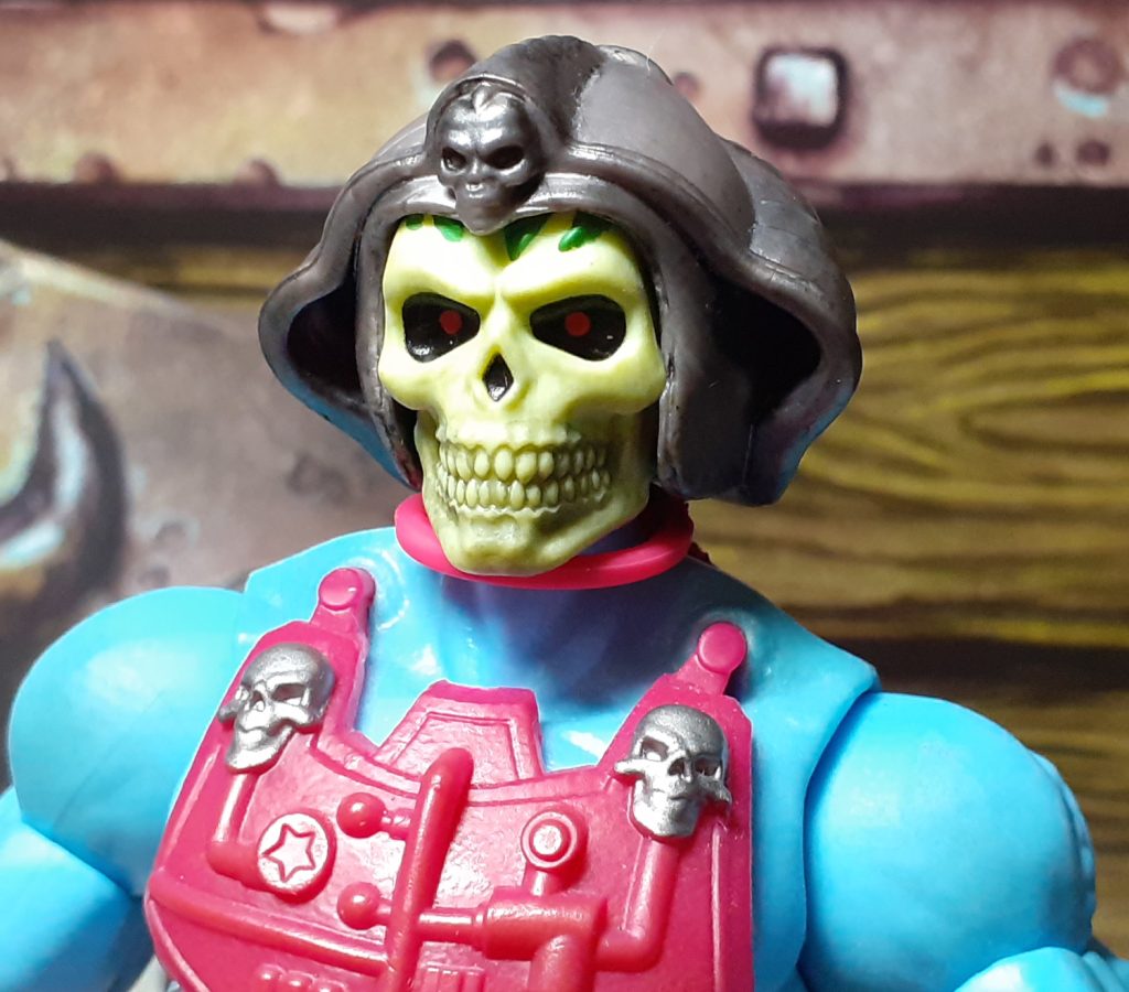 A head and shoulders photo of the New Adventures Skeletor. The figure's shoulders are a light blue and the top of the magenta chest armour with the two silver skulls at the top can be seen. This Skeletor has a large yellow skull face with black eye sockets and dots of red for eyes. It is wearing a gun metal grey helmet which has a curved design with another skull at the top above the forehead.