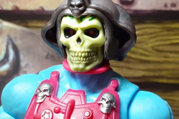 A head and shoulders photo of the New Adventures Skeletor. The figure's shoulders are a light blue and the top of the magenta chest armour with the two silver skulls at the top can be seen. This Skeletor has a large yellow skull face with black eye sockets and dots of red for eyes. It is wearing a gun metal grey helmet which has a curved design with another skull at the top above the forehead.