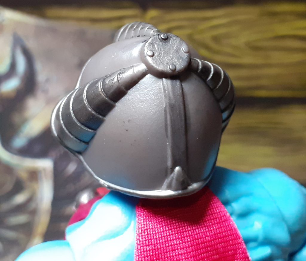 A photo of the back of the Skeletor figure's helmet. The helmet is a gun metal grey with a darker wash picking out some details of the design. It has ridges that meet up at the top of the helmet where there is a central circular point with four small raised pieces that look a little like bolts.