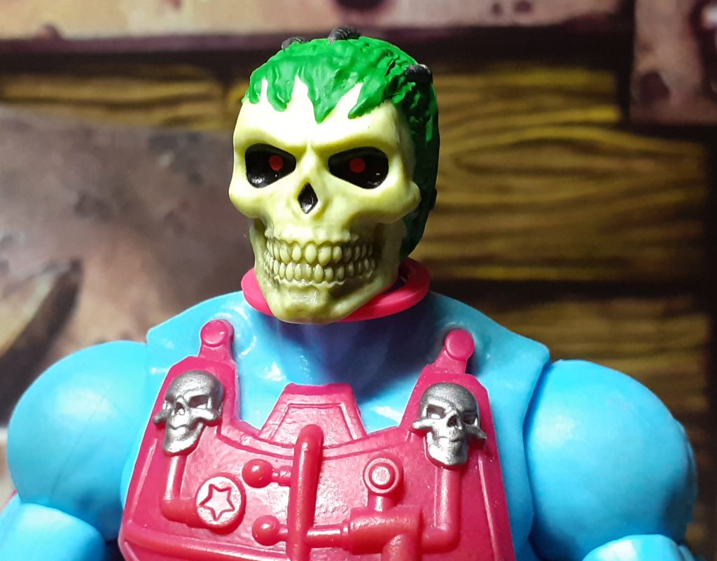 The skull face of this Skeletor without the helmet on. The skull face is yellow with a wash over that picks out details like the teeth. The eye sockets are black and both have red dots for the eyes. Around the top of the forehead is some ridges skin painted a sickly looking green.