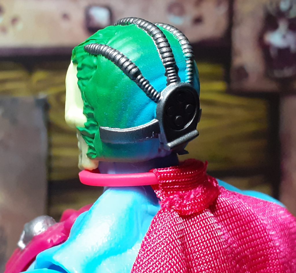 A photo of the back of the figure's head. From the front, the sickly looking green skin fades into a light blue colour. On the back of the head is a piece of silver coloured cybernetic implant that has tubes spreading out from it over the top of the head.