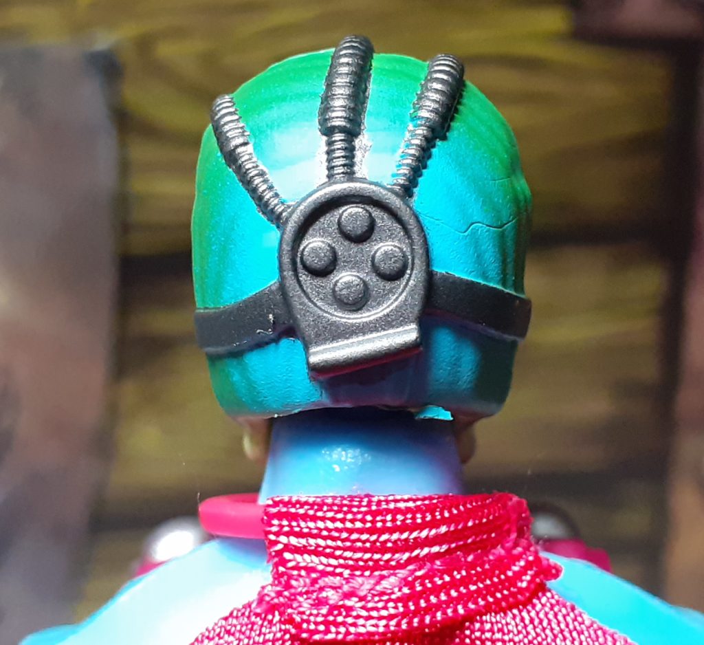 Another photo of the back of the head, this time looking straight on at it. The silver cybernetic implant is more clear. It has a roughly circular design with four small round parts in the middle. These look like they could be the visible parts of bolts or screws. On either side of this piece are straps holding it onto the head. From the top extend tubes that go over the head towards the face.