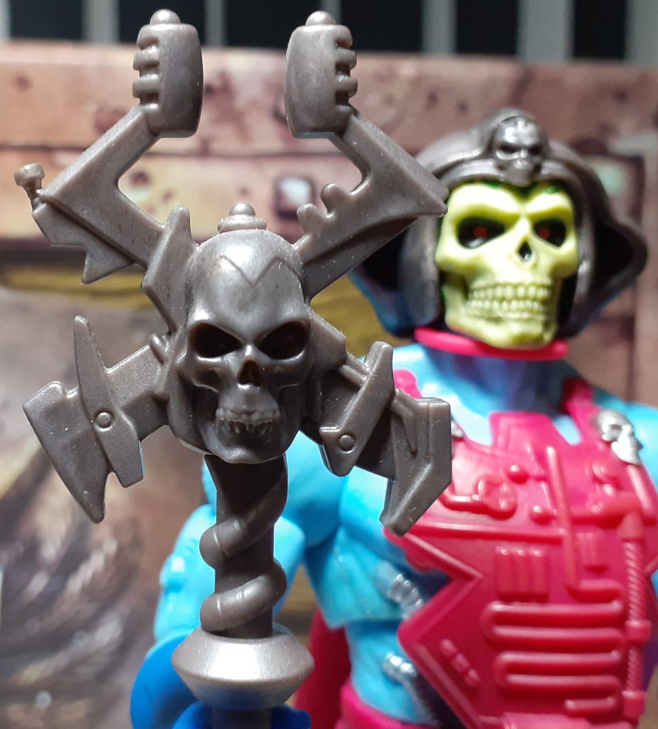 A close up photo of the top of the Havoc Staff. It's moulded in a gen metal grey plastic with a darker wash over it. The central part has a human skull with four protrusions coming out of it. The bottom two look like blades while the top two look a bit like shock prods.