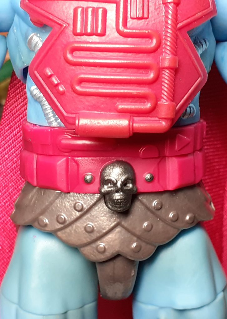 A close up photo of the New Adventures Skeletor figure's midriff. The bottom half of the torso is mostly covered by a magenta coloured piece of chest armour. The bottom of this overlaps the large belt, also magenta in colour. Under this are the figure's metallic looking trunks with a skull on the front!