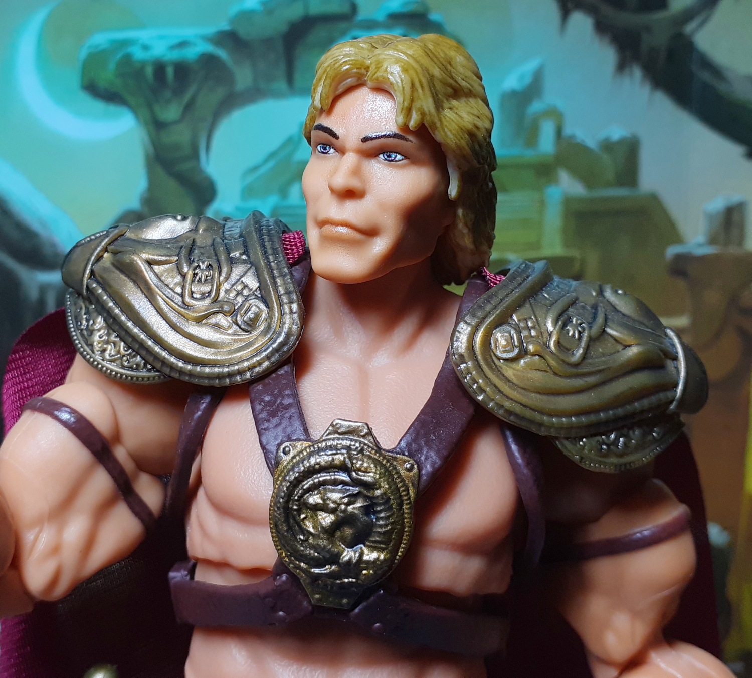 A photo of the upper half of the 1987 Movie He-Man figure. The figure is wearing armour which is comprised of shoulder pauldrons in a dark gold colour with leather looking straps across the chest. In the centre of the chest is a dragon emblem in more of the dark gold coloured plastic.