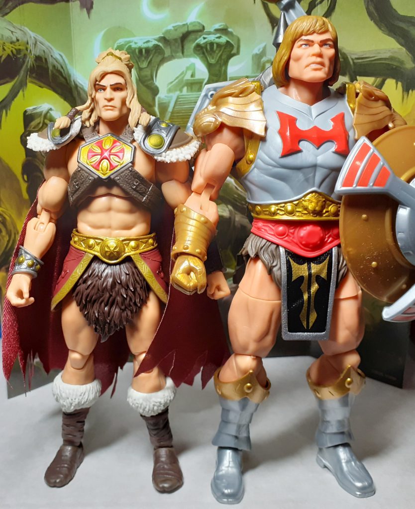 A side by side height comparison photo of the King Grayskull and Flying Fists He-Man figures. On the left of the photo is King Grayskull while on the right is Flying Fists He-Man. This version of He-Man has gold and silver armour with lion pauldrons over the shoulders.