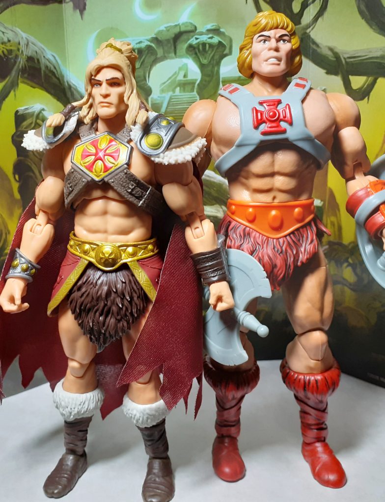 A side by side height comparison photo of the King Grayskull and 40th Anniversary He-Man figures. On the left of the photo is King Grayskull while on the right is this version of He-Man. This He-Man is wearing the classic grey chest harness with red cross but also has an overlarge and muscular torso.