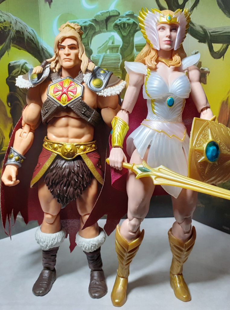 A side by side height comparison photo of the King Grayskull and New Etheria She-Ra figures. On the left of the photo is King Grayskull while on the right is She-Ra. This version of She-Ra has a white and gold head dress with a winged design over her head.