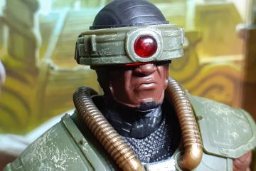 A head and shoulders photo of Tri-Klops looking towards the right of the photo. Tri-Klops is a three-eyed warrior with the eyes mounted on a rotating visor. In this photo the circular eye is facing forwards and has a ruby red lens in it. The armour is green over a dark brown or black futuristic looking kevlar suit. Over each shoulder is a gold coloured tube too.